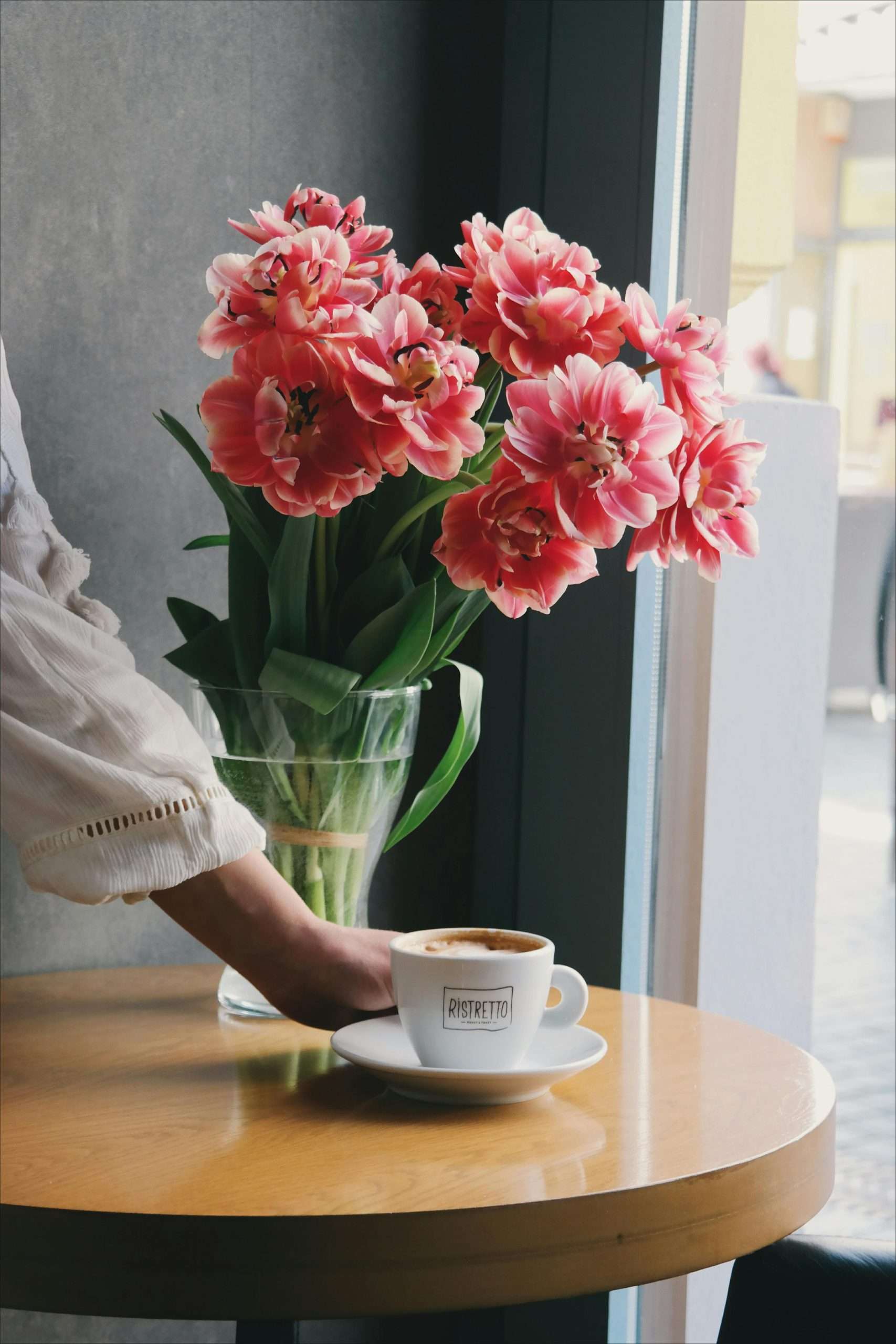 person placing a cup of latte on a white saucer near pink flowers in bloom