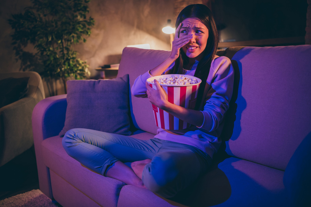 Photo of homey chinese lady eat popcorn movie night watch horror, scary movie hiding eyes afraid look on blood sit cosy couch casual outfit dark living room indoors